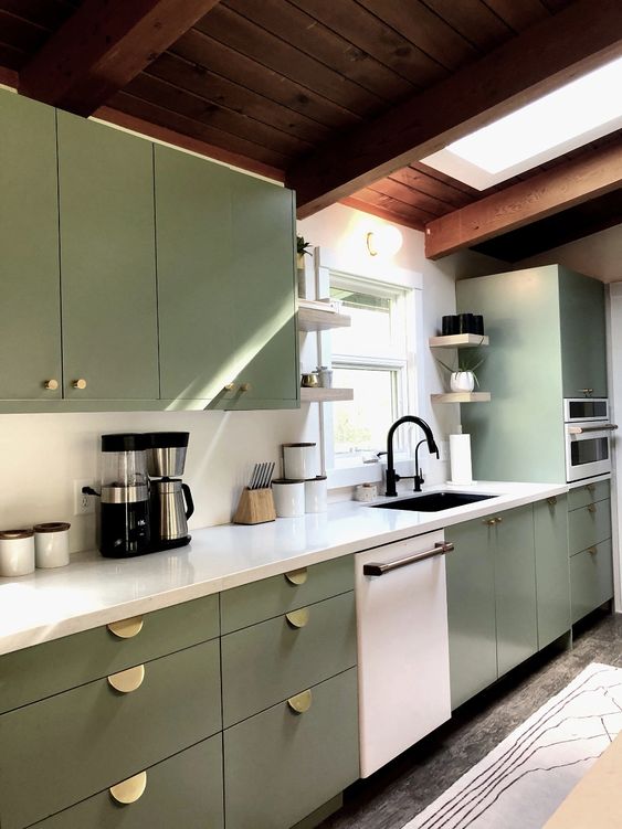 a sage green flat panel kitchen with gold pulls, a white backsplash and countertops, black fixtures and a skylight over the space