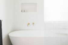 a serene neutral bathroom with a terrazzo floor, skinny tiles, an oval tub and a fluted glass shower screen