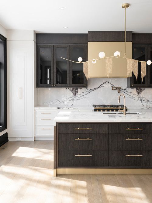 a sophisticated dark and white kitchen with flat panel cabinets, glass front ones, a white marble backsplash, a gold hood and a catchy lamp
