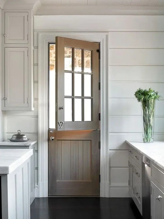 a stained wood Dutch door adds a rustic feel to the all-white kitchen with paneling and makes it more welcoming