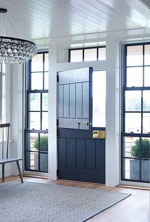a stylish modern entryway with a navy Dutch door and windows on both sides, with a wooden bench, a refined chandelier and a simple rug