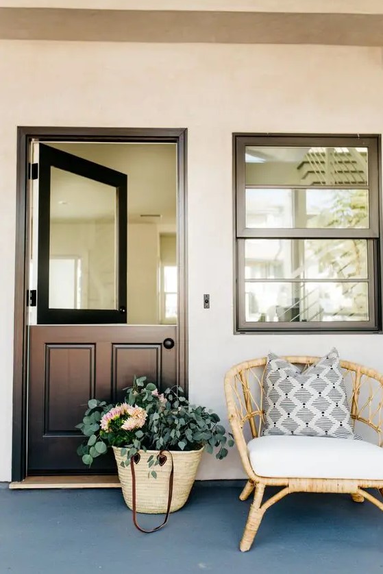 a stylish modern meets rustic entrance with a black Dutch door with a solid glass insert and a rattan chair plus greenery in a woven bag