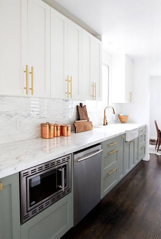 a trendy two-tone kitchen with white and sage green cabinets, a white quartz backsplash and countertops and gold handles