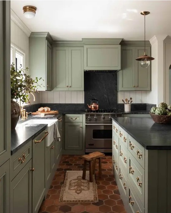 a vintage green kitchen with shaker style cabinets, black countertops and a backsplash, gold and brass fixtures and pendant lamps