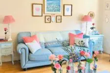 a vintage living room with light yellow walls, a blue sofa and pink pillows, a pink upholstered ottoman, a gallery wall and pink lamps