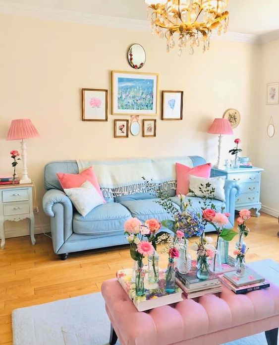 a vintage living room with light yellow walls, a blue sofa and pink pillows, a pink upholstered ottoman, a gallery wall and pink lamps