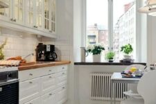 a welcoming white Scandinavian kitchen with glass cabinets, white tiles, butcherblock countertops and a small eating zone