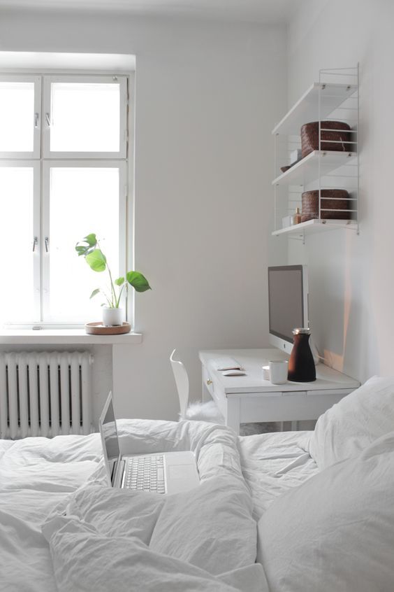 a white Scandinavian bedroom with a bed with white bedding, a desk and a chair, an open shelf and some greenery