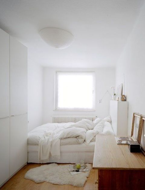 a white bedroom with a bed, a storage headboard, a wardrobe, a stained desk, white textiles and artwork