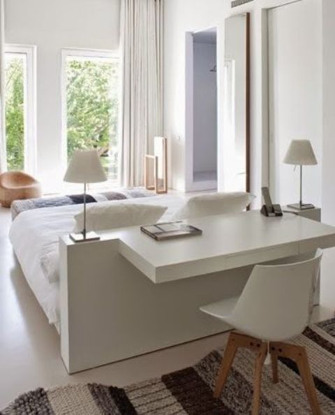 an airy white bedroom with a bed that features a white desk built-in, a white chair, a woven one and white lamps