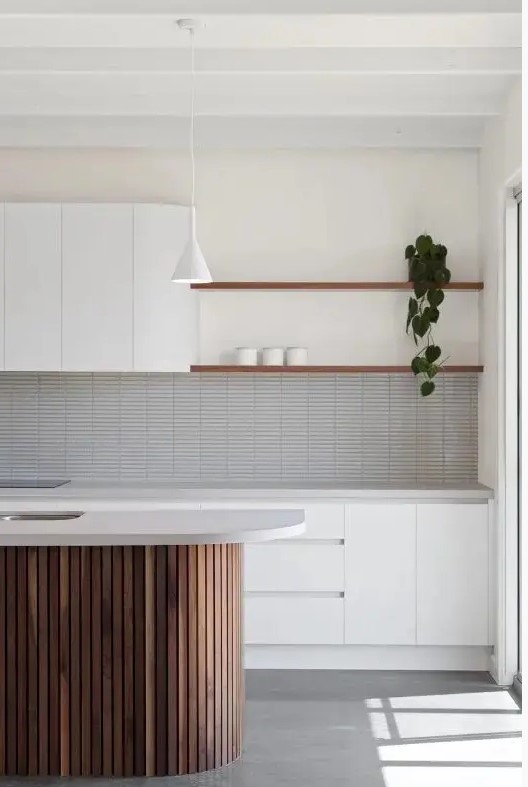 an airy white kitchen with plain cabinets and no handles, a grey skinny tile backsplash, a fluted stained kitchen island and shelves