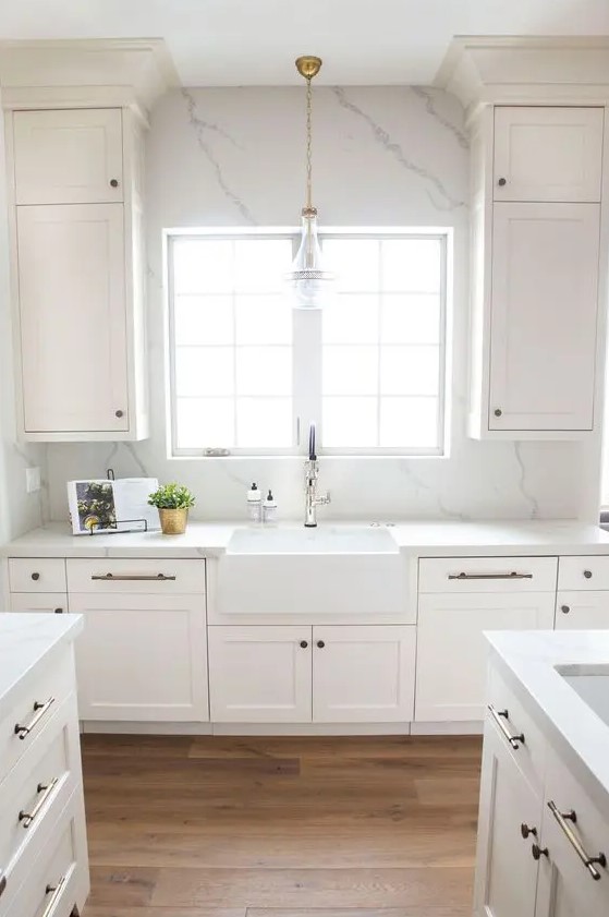 an airy white kitchen with shaker cabinets, a white quartz backsplash and countertops plus brass fixtures is wow