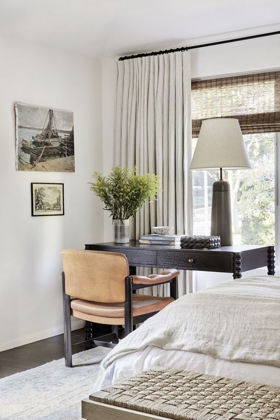 an elegant neutral bedroom with a bed with neutral bedding, a black desk, a leather chair, artwork and some greenery