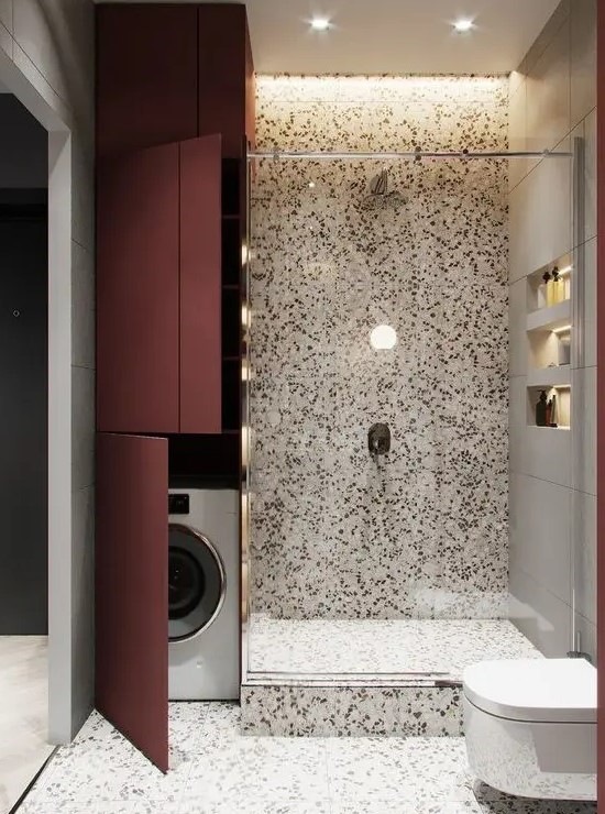 a bright bathroom done with terrazzo with a washing machine hidden by the shower space and behind burgundy doors