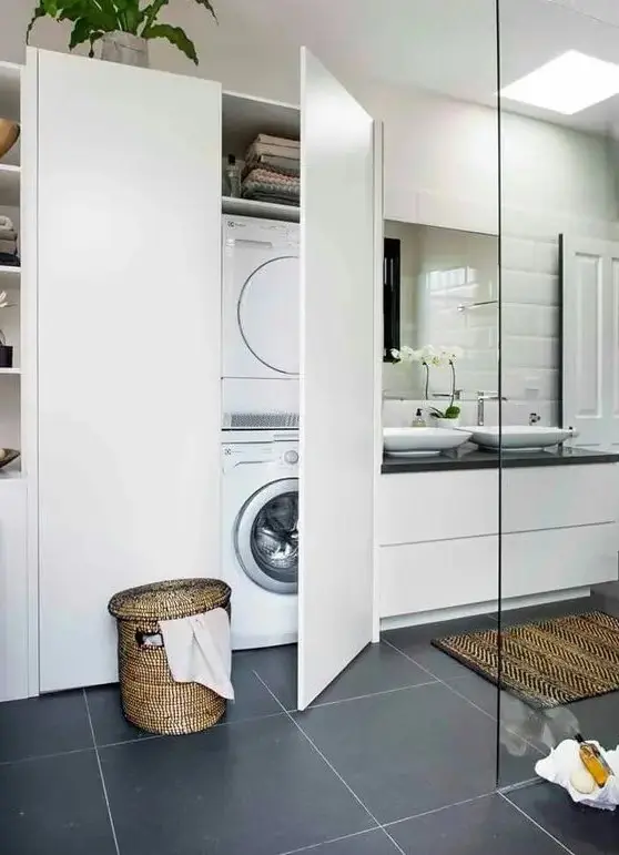 a modern bathroom clad with grey and white tiles, a white vanity with sinks, a large storage unit with a washing machine hidden