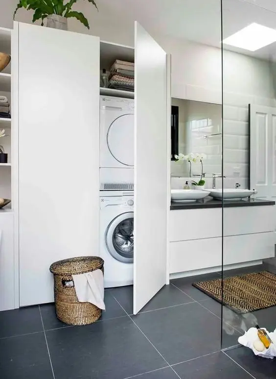 a modern white bathroom clad with large scale tiles, a white vanity with a black countertop, a large white storage unit with a washing machine and a dryer