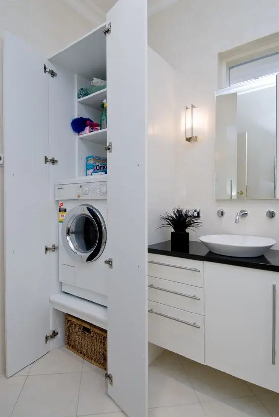 a small white bathroom with a built in vanity, a mirror, a small storage cabinet with a washing machine and sconces