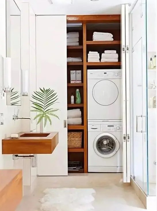 a white bathroom with a glass enclosed shower space, a large storage unit, a washing machine and a dryer and shelves for storage, a wooden sink