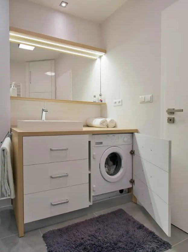 a white bathroom with plain walls, a mirror with a shelf, a vanity with drawers and a small washing machine in the vanity