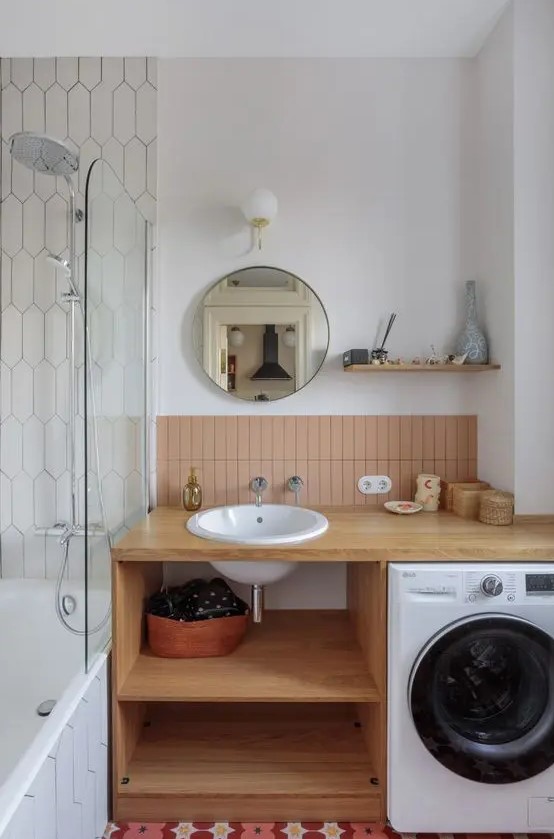 a mid century modern bathroom with a bathtub, an open timber vanity with a washing machine, dusty pink tiles and a round mirror