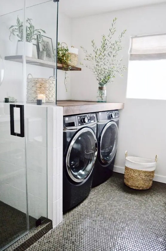 a modern bathroom with penny tiles, a glass enclosed shower, a washing machine and a dryer, greenery and a basket