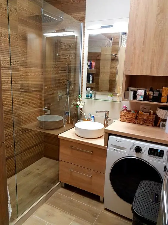 a small and cozy wood clad bathroom with a shower space, a small vanity, a washing machine and a storage unit, a lit up mirror