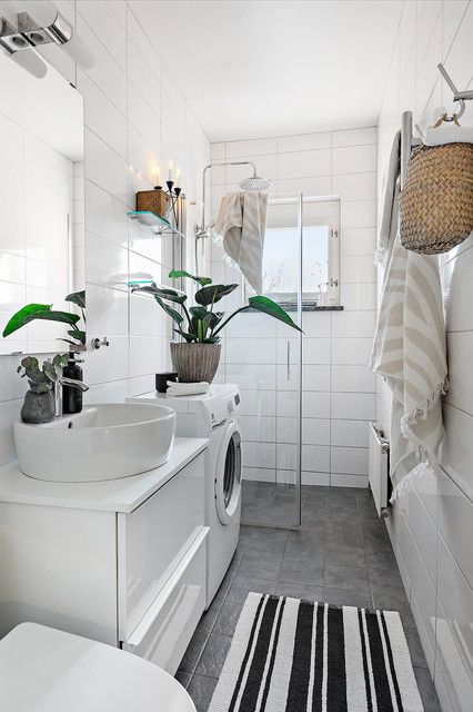 a small bathroom clad with large scale tiles, with a shower space, a washing machine and a white vanity, potted plants and baskets