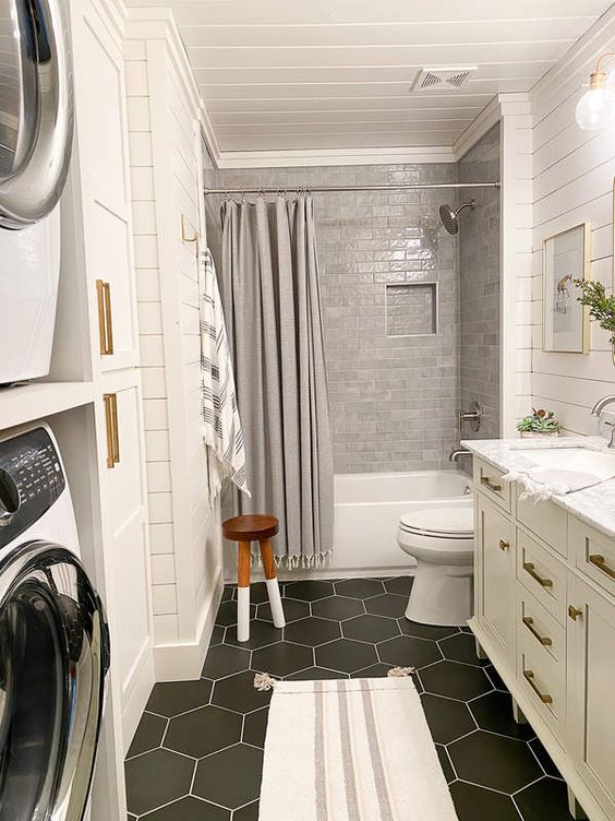 a small farmhouse bathroom with a black hex tile floor, a shower space clad with grey tiles, a large vanity and a stacked washing machine and dryer