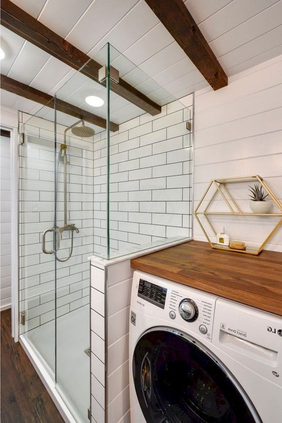 a white farmhouse bathroom clad with beadboard and white subway tiles, a washing machine with a butcherblock countertop, a shower space and wooden beams