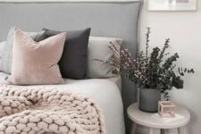 a Scandinavian bedroom with a grey upholstered bed, pink and grey pillows and a pink knit blanket, a grey planter with greenery