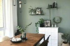 a Scandinavian dining room with a sage green accent wall, a rustic table, black and white chairs, pendant bulbs and open shelves