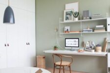 a Scandinavian home office with a sage green accent wall, a wall shelving unit and a desk, a round table and a white chair