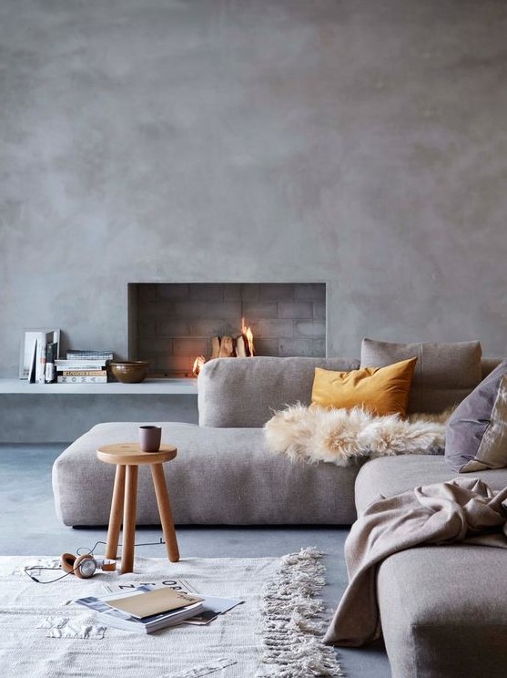 a Scandinavian living room with concrete walls and a floor, a grey sectional with pillows, a wooden stool and some books is cool