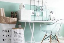 a Scandinavian workspace with a mint green accent on the wall, a desk and a clear chair, some paper bags and a memo board