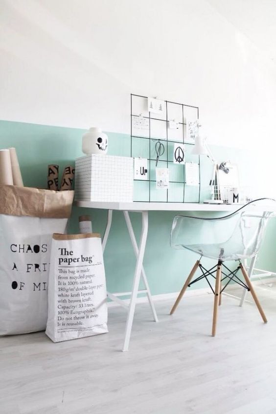 a Scandinavian workspace with a mint green accent on the wall, a desk and a clear chair, some paper bags and a memo board