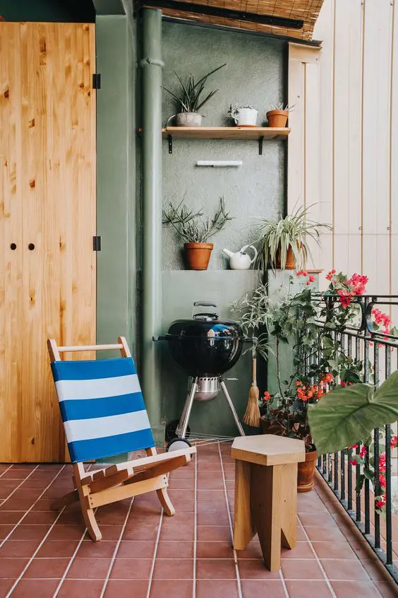 a balcony done with green walls, a terracotta tile floor, a folding chair, a stool, a grill and lots of plants and blooms in pots around