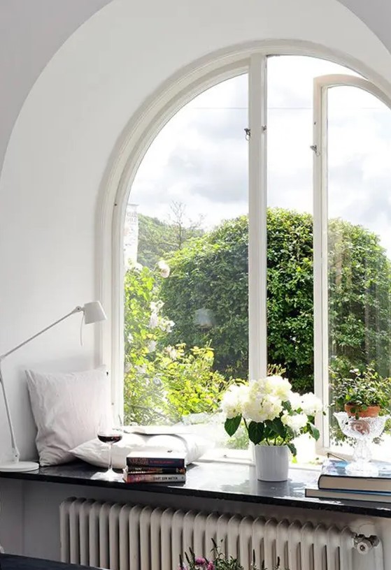 a beautiful arched casement window, a windowsill styled with pillows, blooms and greenery and book stacks are very cozy and chic