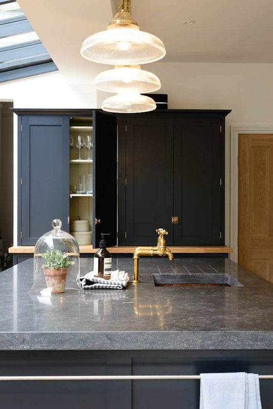 a beautiful black kitchen with shaker cabinets and a grey soapstone countertop plus gold fixtures and pendant lamps