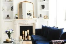 a beautiful neutral living room with a candle fireplace, a navy sofa, built-in shelves, a glass coffee table and lovely gold touches here and there