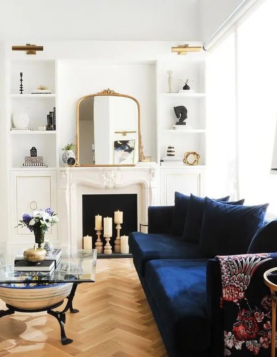 a beautiful neutral living room with a candle fireplace, a navy sofa, built in shelves, a glass coffee table and lovely gold touches here and there