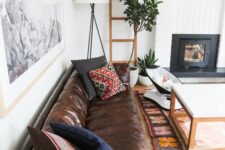 a boho living room with a built-in fireplace, a brown leather sofa, printed pillows, potted plants and a large artwork