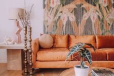 a boho living room with a floral tapestry, an orange sofa, a round table and a side table plus candle holders and a boho rug