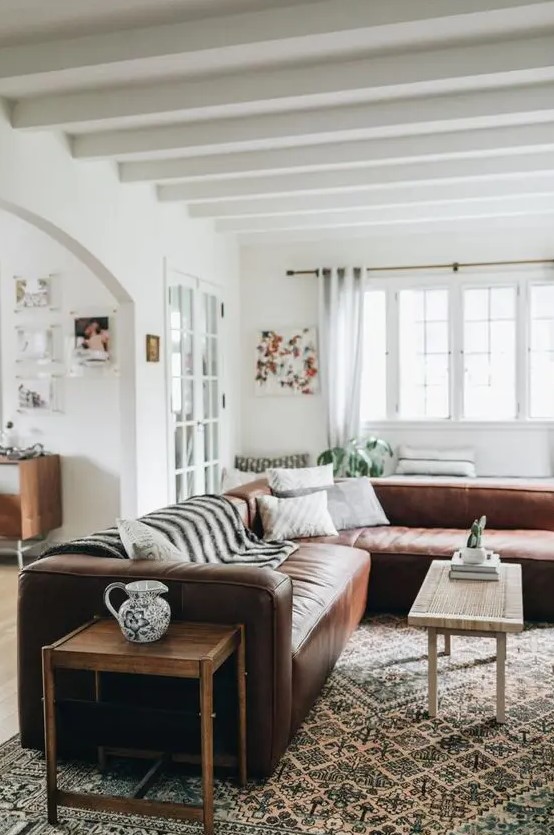 a boho rustic interior with a brown leather sectional, a coffee table and a side one, a large printed rug and pillows