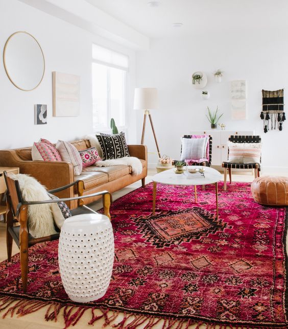 a bold boho living room with a tan leather couch, a bright pink printed rug, some chairs, a coffee table and a leather pouf