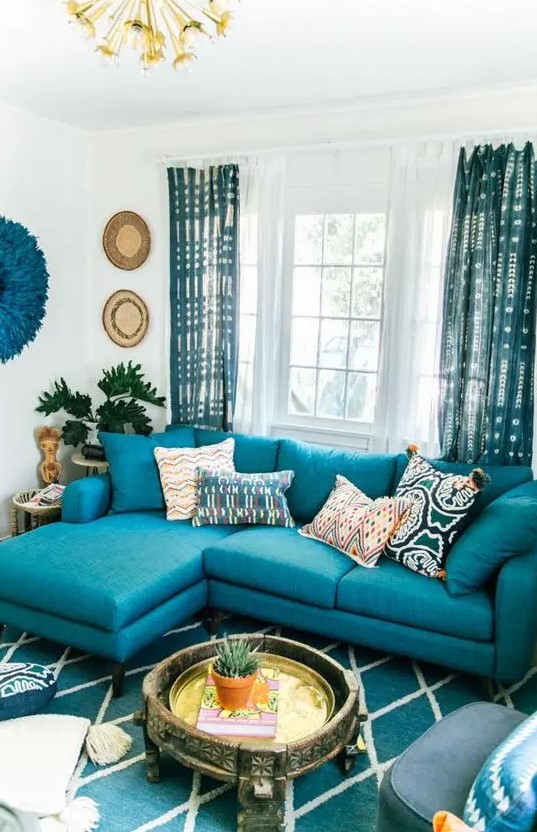 a bright living room with a turquoise sofa, a turquoise printed rug and curtains, decor on the wall and a carved wooden coffee table
