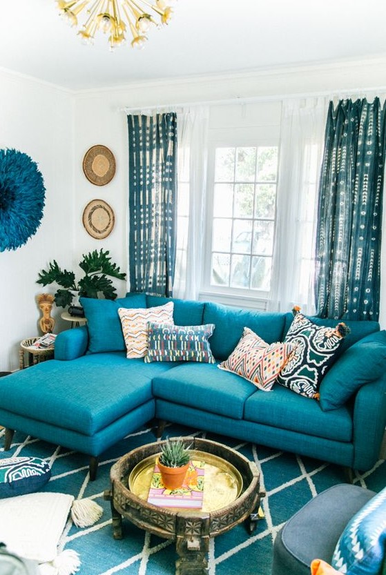a bright living room with a turquoise sofa, a turquoise printed rug and curtains, decor on the wall and a carved wooden coffee table