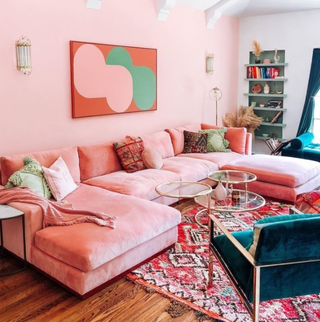 a bright living room with pink walls, a pink sofa, colorful pillows, teal chairs, glass tables, built in shelves and teal curtains