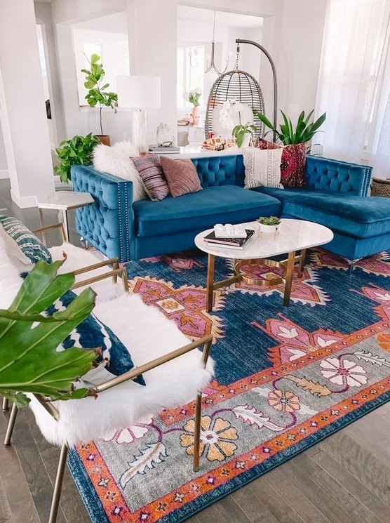 a bright mid-century modern living room with a bold printed rug, a blue sofa, white faux fur chairs, an oval coffee table and greenery