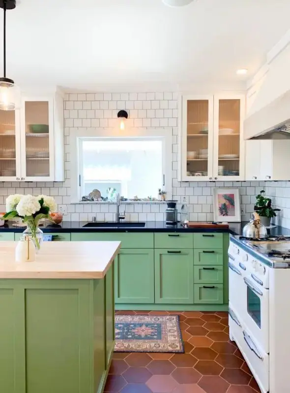 a bright modern kitchen with white fluted glass and green shaker style cabinets, black countertops, white square tile walls and a terracotta tile floor