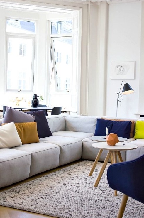 a bright modern living room with a grey low sofa, a bold blue chair, colorful pillows, some lamps and round tables